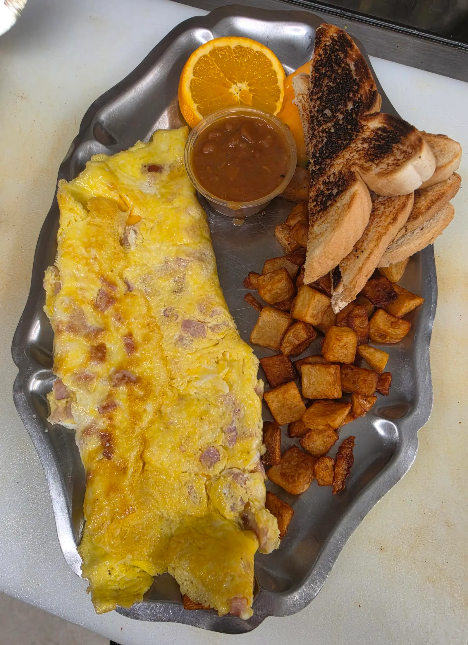Omelette au jambon servie avec patates en cubes rissolées, pain grillé, fèves en sauce et une tranche d'orange
