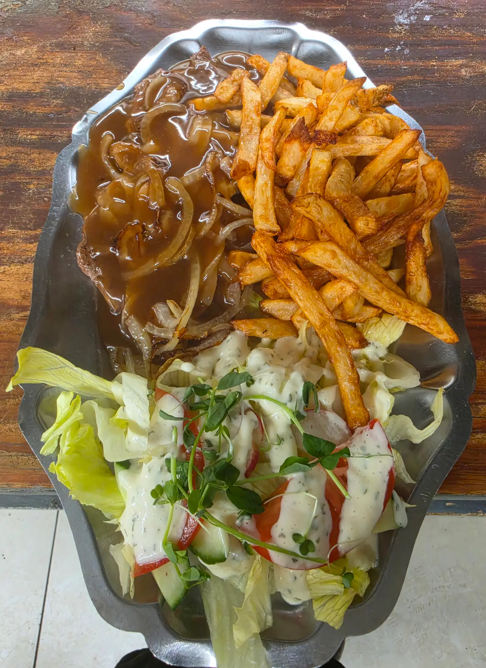 Pièce de viande nappée de sauce brune et d'oignons, avec frites et salade garnie de pousses, sur un plat en métal